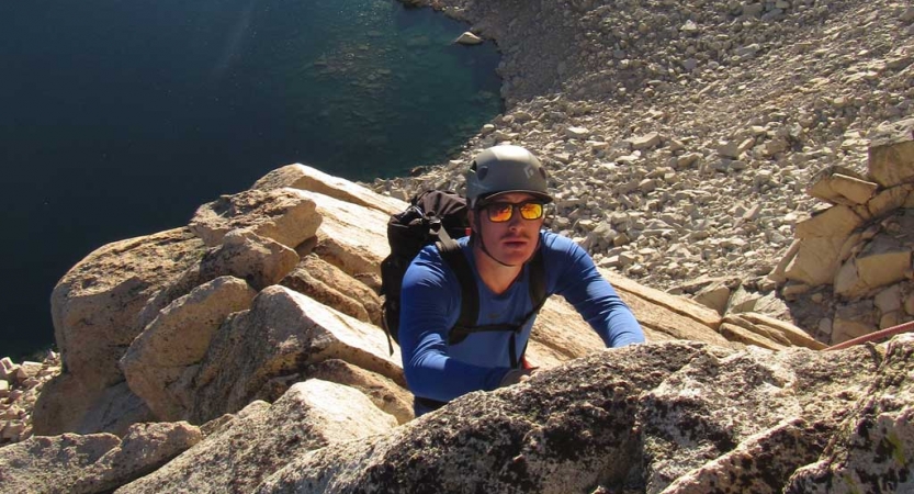 A person looks up at the camera while rock climbing on an outward bound gap year course. There is a vast rocky landscape and an alpine lake below them. 